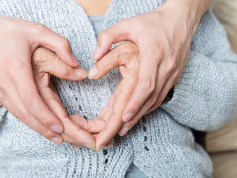 Two pairs of hands, one older and one younger, gently form a heart shape together against the chest of a person wearing a light gray knitted sweater, symbolizing care, love, and connection across generations.