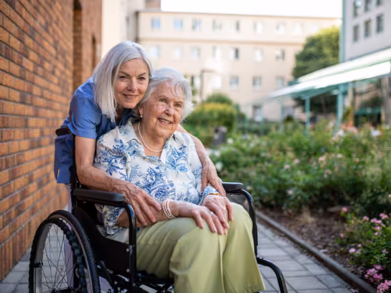 An elderly woman sits in a wheelchair, smiling, while a younger woman stands behind her, hugging her shoulders. They are outdoors on a sunny day, in a garden area next to a building.