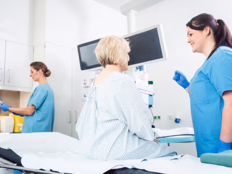 A patient in a hospital gown sits on an exam bed, facing a smiling healthcare worker in scrubs and gloves. Another staff member is working in the background by a counter in a bright medical room.