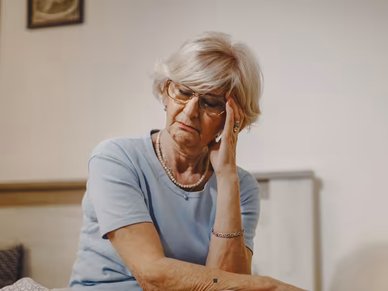 An elderly woman with gray hair and glasses sits on a bed, resting her head on her hand. She appears thoughtful or worried, wearing a light blue shirt and pearl necklace, in a softly lit room.