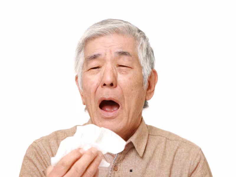 An older man with gray hair is sneezing or about to sneeze, holding a tissue in one hand, with his eyes closed and mouth open, against a plain white background.