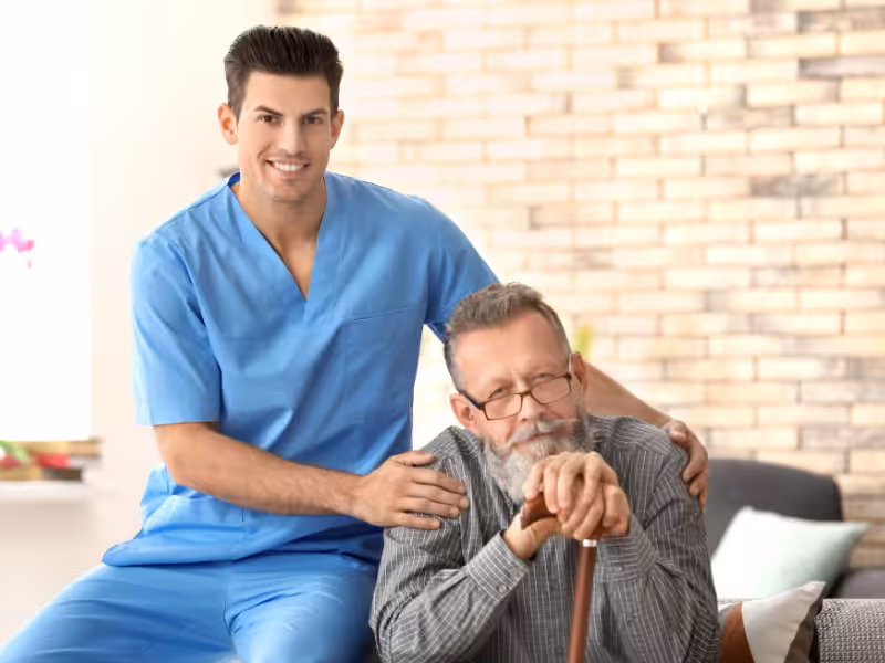 A young male caregiver in blue scrubs smiles while gently resting his hand on an elderly man with glasses and a gray beard, who is seated and holding a cane in a cozy, well-lit room.