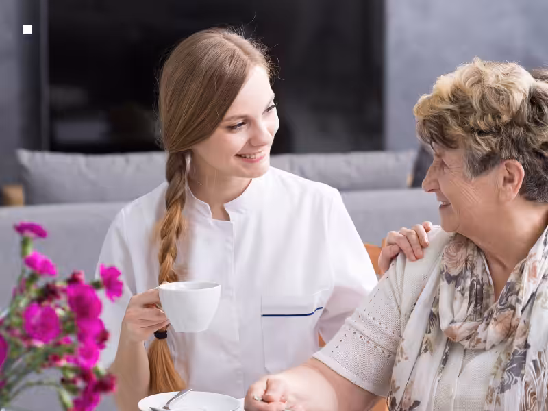 A young caregiver with a long braid smiles warmly at an elderly woman, both seated indoors. The caregiver holds a teacup, while the elderly woman wears a scarf. Pink flowers are visible in the foreground.