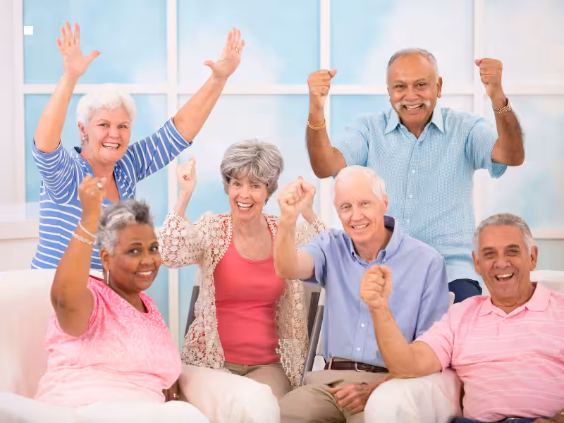 A group of six smiling older adults sit together indoors, raising their fists in celebration and showing excitement. They are dressed in casual, colorful clothing and appear happy and enthusiastic.