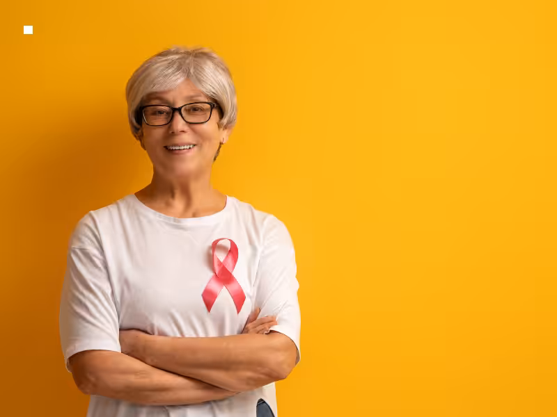 Smiling older woman with short gray hair and glasses stands with arms crossed, wearing a white shirt with a pink breast cancer awareness ribbon, against a yellow background.