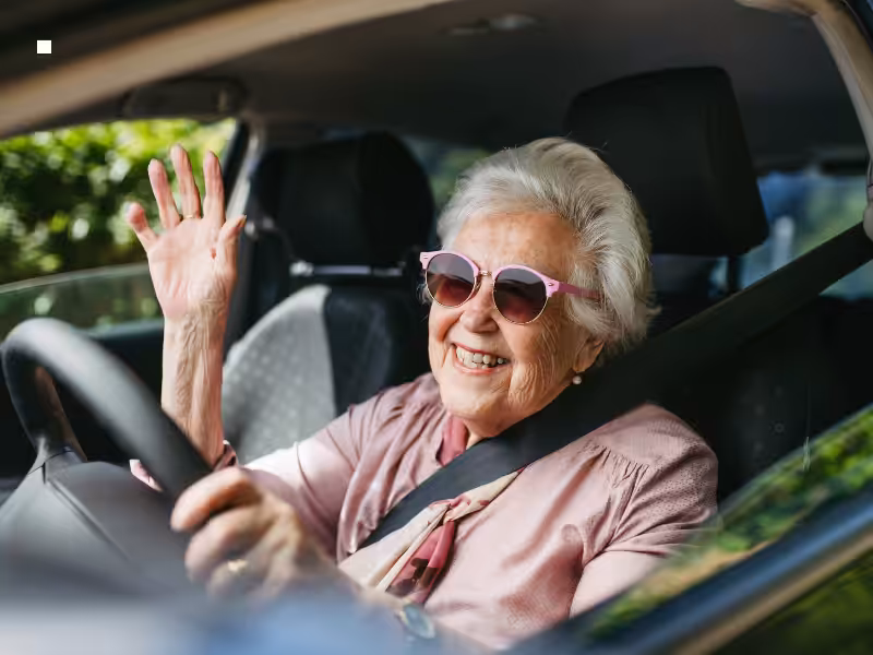 Smiling elderly woman wearing sunglasses waves while sitting in the driver’s seat of a car, wearing a seatbelt and enjoying a sunny day.