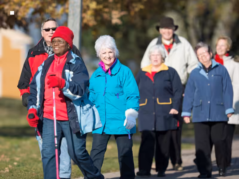 A group of older adults wearing colorful jackets walk together outdoors on a sunny day, smiling and enjoying each others company. Trees with autumn leaves are visible in the background.