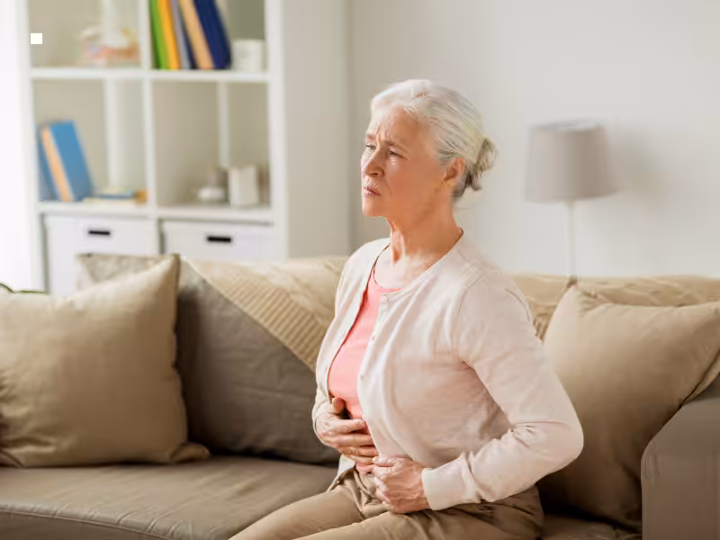 An older woman with gray hair sits on a beige sofa, holding her stomach with both hands and appearing to be in pain or discomfort. Shelves and a lamp are visible in the background.