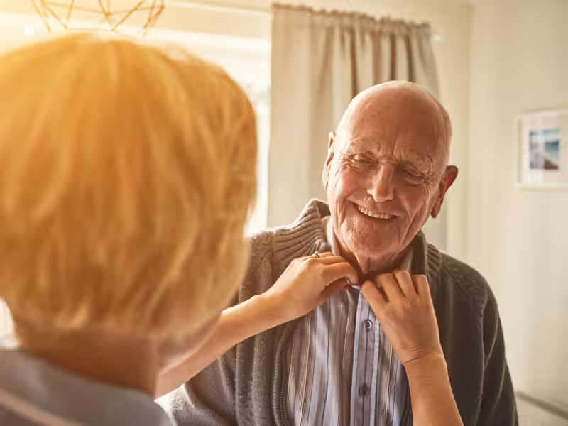 A caregiver is helping an elderly man button his shirt in a warmly lit room. The man is smiling, and they appear to be enjoying a pleasant, caring moment together.