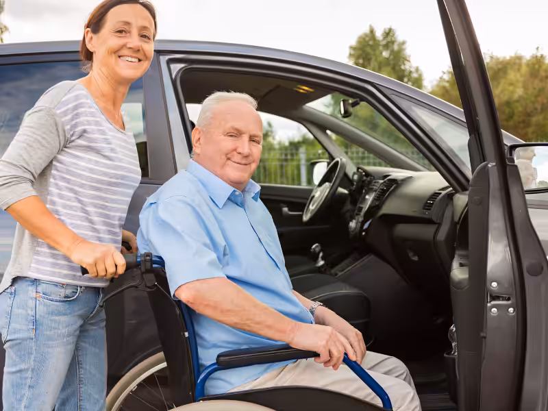 A smiling older man in a wheelchair is being helped into a parked car by a woman standing beside him, both looking at the camera. The car door is open and it is daytime.