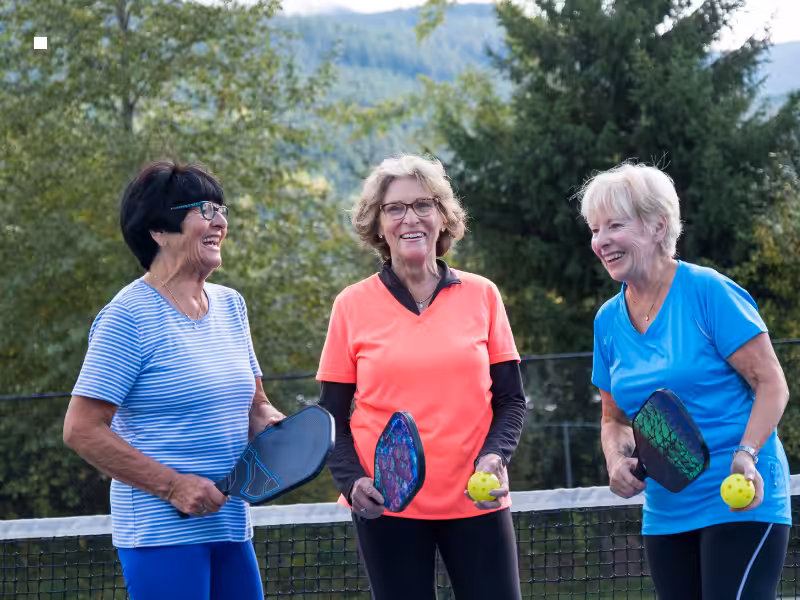 Three older women stand outdoors on a pickleball court, smiling and holding paddles and yellow balls. They are dressed in sporty clothing with trees and hills in the background.