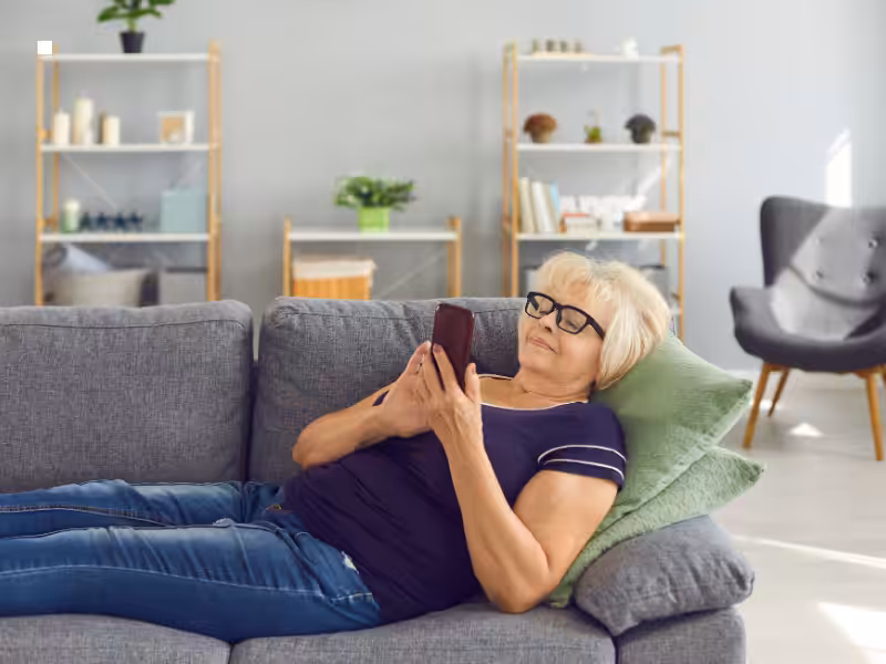 Older woman wearing glasses lies on a gray couch, propped up by a green pillow, smiling while using her smartphone. The living room has modern decor, shelving, and plants in the background.