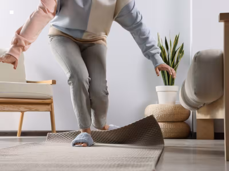 A person in slippers is tripping over a bunched-up rug in a living room, with one arm outstretched for balance. A plant and cozy furniture are visible in the background.