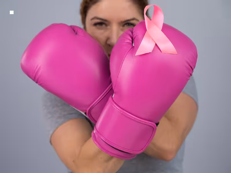 A person wearing pink boxing gloves crosses their arms in front of their face. A pink breast cancer awareness ribbon is on one glove. The background is plain and gray.