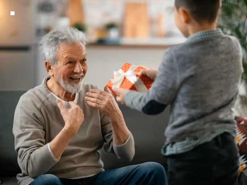 A smiling elderly man sits on a couch, looking delighted as a young boy hands him a red and white polka-dotted gift box with a ribbon. The setting appears warm and homey.