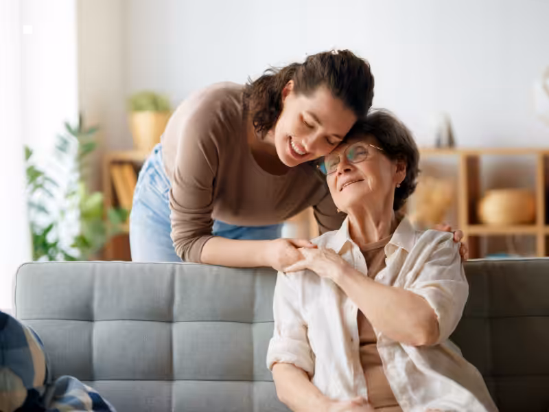A young woman leans over a couch to hug an elderly woman, both smiling warmly in a cozy, well-lit living room.