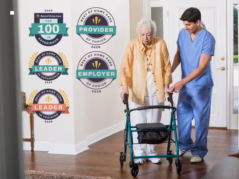 A caregiver in blue scrubs assists an elderly woman using a walker at home. Next to them are five award badges for home care, highlighting experience, training, provider, and employer recognition.