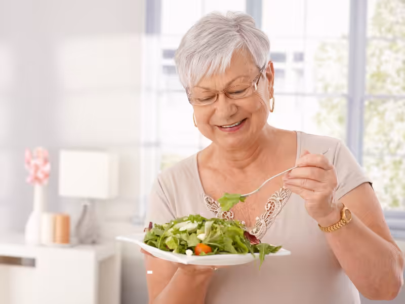 Older woman with short gray hair and glasses smiling while holding a plate of salad and eating with a fork in a bright, modern kitchen.