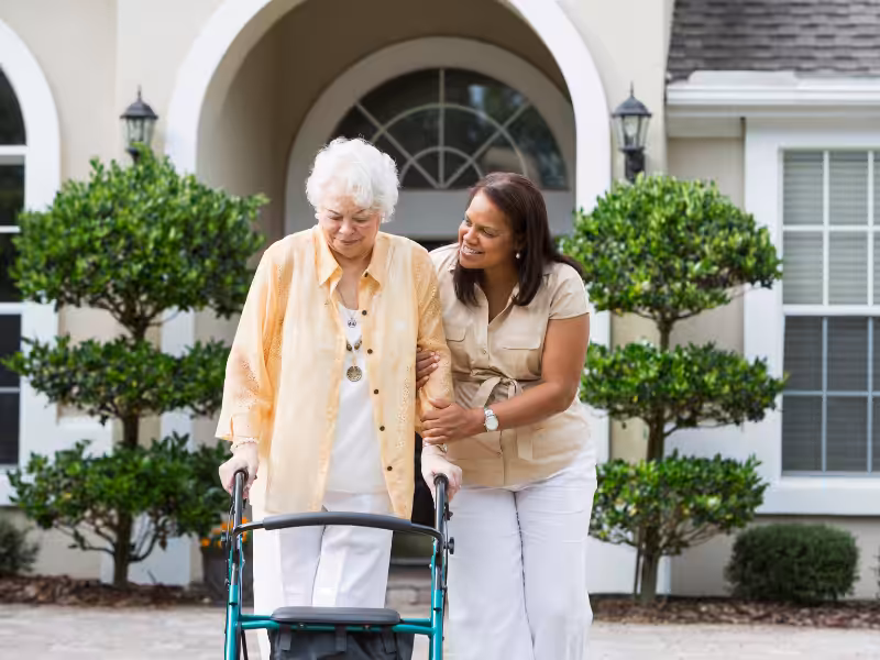 An elderly woman using a walker is assisted by a younger woman outside a house with arched windows and neatly trimmed bushes. Both women are smiling and appear to enjoy each other’s company.