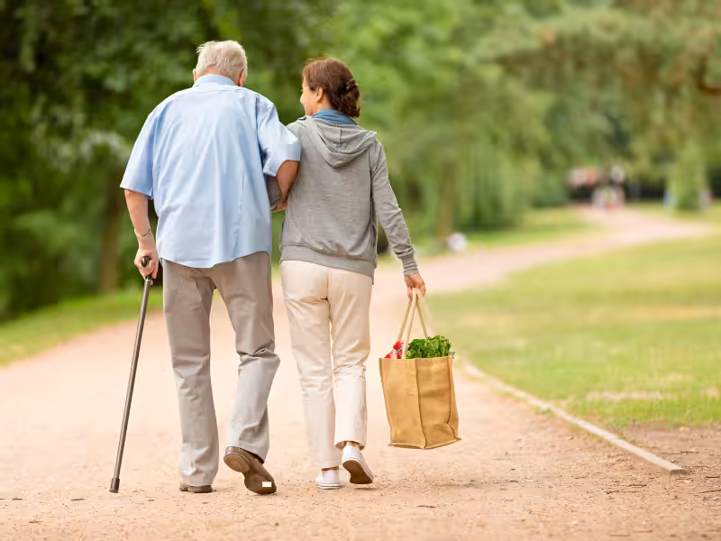 An elderly man with a cane walks arm-in-arm with a younger woman carrying a grocery bag along a tree-lined path in a park.