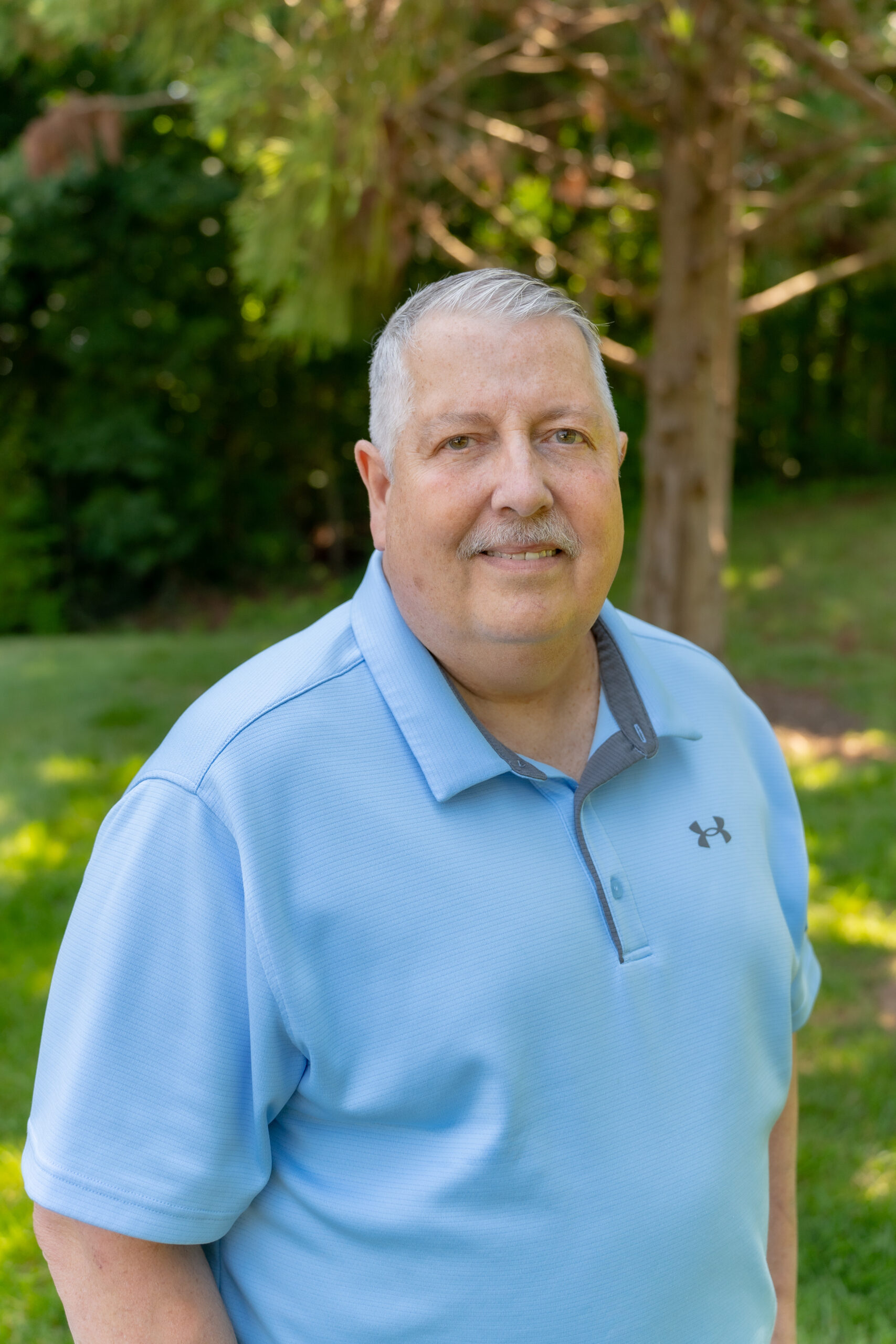 A man with short gray hair and a mustache, wearing a light blue Under Armour polo shirt, stands outside on a sunny day with trees and green grass in the background.