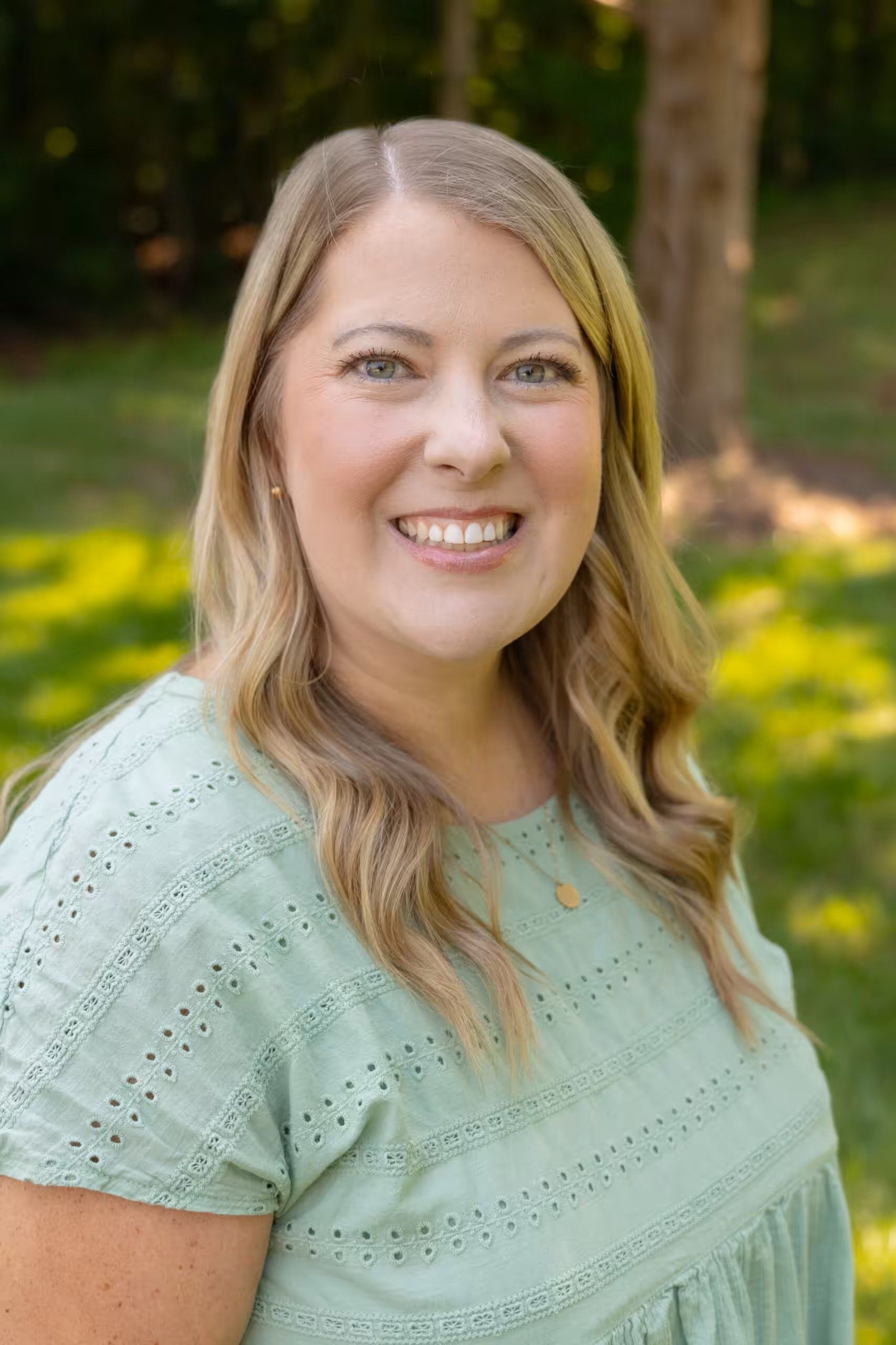 A woman with long blonde hair wearing a light green eyelet blouse smiles outdoors, with a grassy background and trees in soft focus behind her.