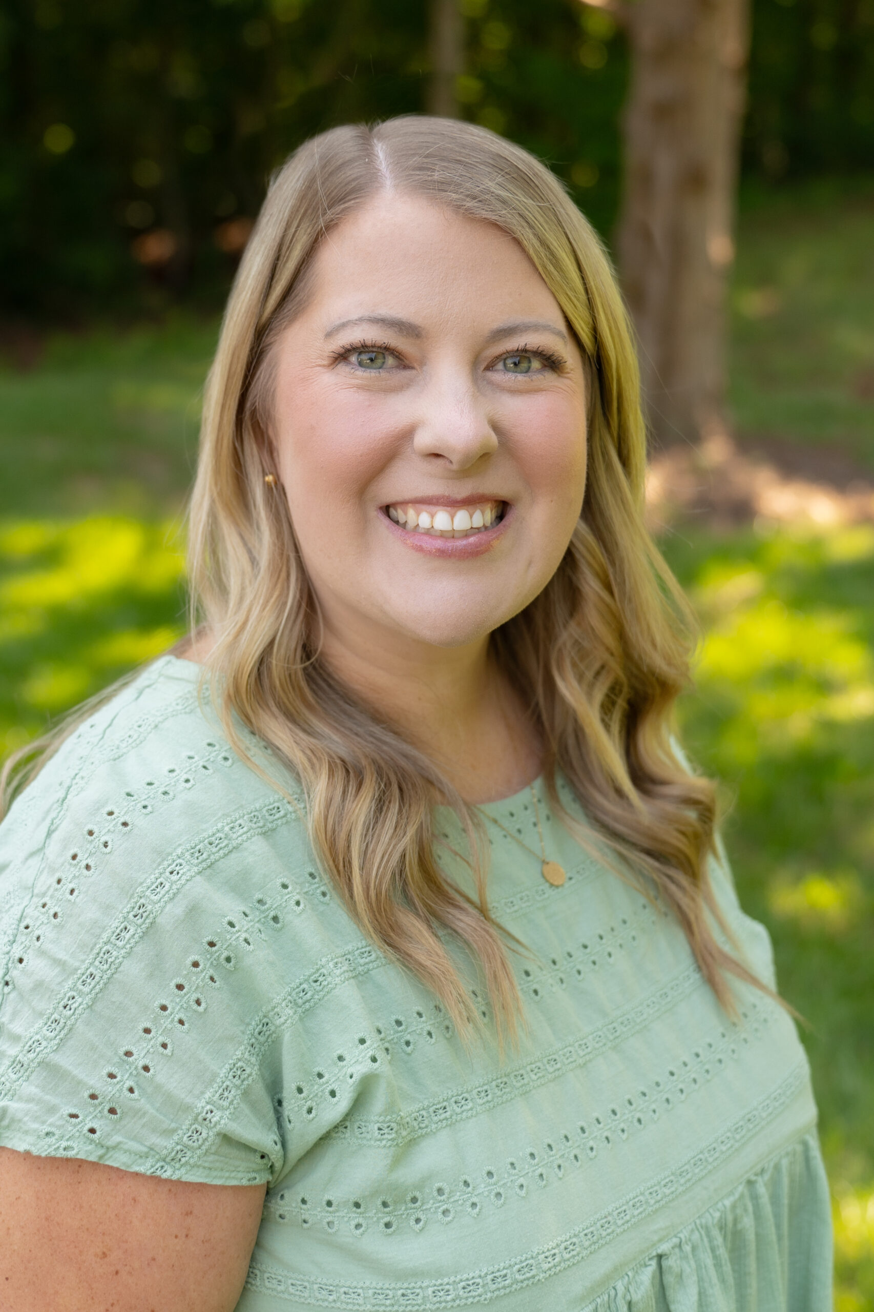 A woman with long blonde hair wearing a light green eyelet blouse smiles outdoors, with a grassy background and trees in soft focus behind her.