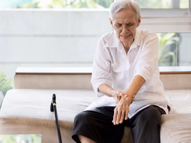 An elderly person with gray hair, wearing a white shirt and black pants, sits on a sofa holding their painful arm, with a cane resting nearby.