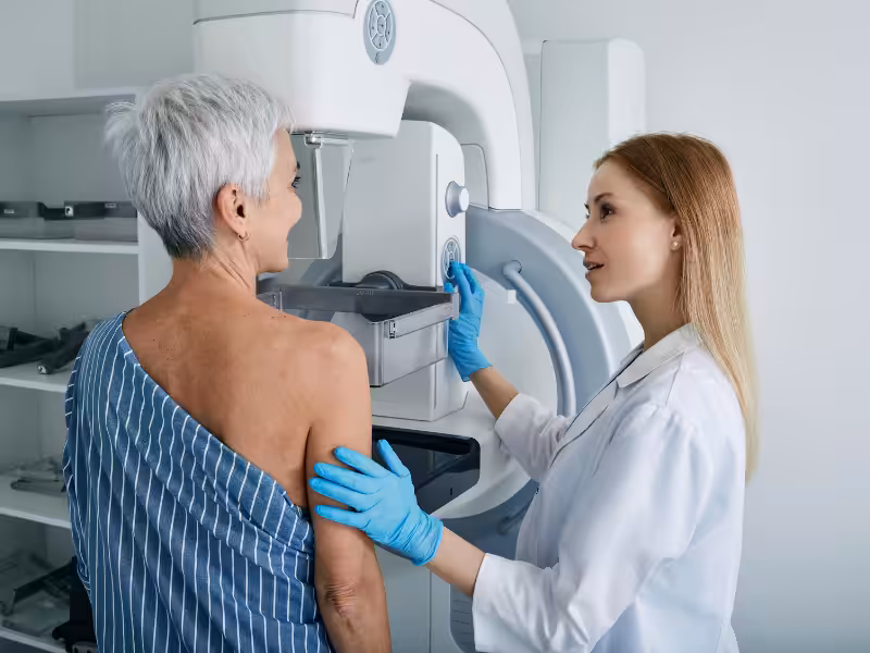 A healthcare professional in a white coat and blue gloves assists an older woman with short gray hair during a mammogram procedure in a medical office.