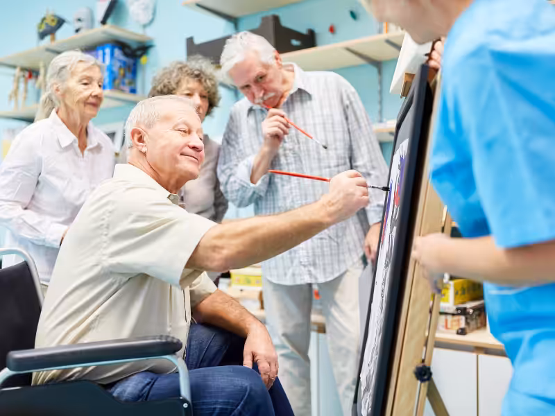 A group of older adults, some standing and one in a wheelchair, smile and paint together on a canvas in an art class, guided by an instructor in blue scrubs.