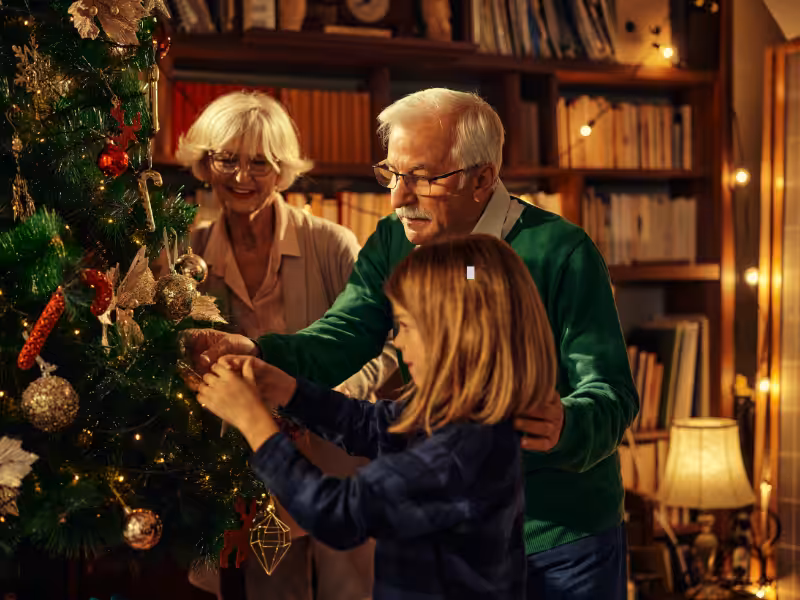 An elderly couple and a child decorate a Christmas tree together in a warmly lit, cozy room filled with books and festive ornaments.