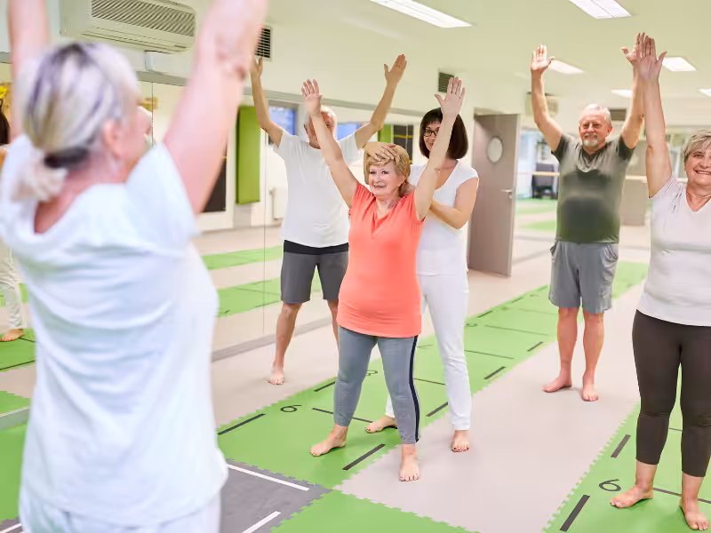 A group of older adults participate in a fitness class, standing on mats with arms raised, following the instructor at the front of the room in a bright, spacious studio.