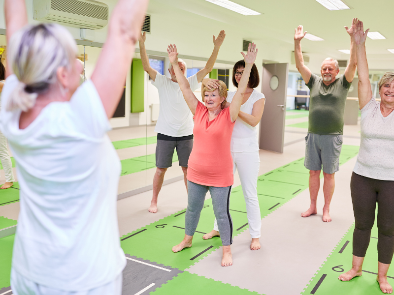 A group of older adults participate in a fitness class, standing on mats with arms raised, following the instructor at the front of the room in a bright, spacious studio.