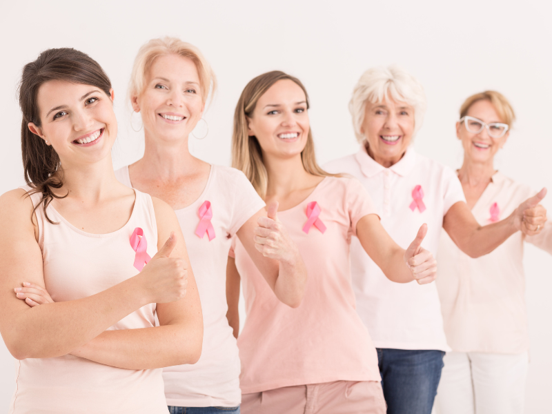 Five women of different ages wearing pink shirts and pink breast cancer awareness ribbons stand in a line, smiling and giving thumbs up, symbolizing support and positivity.