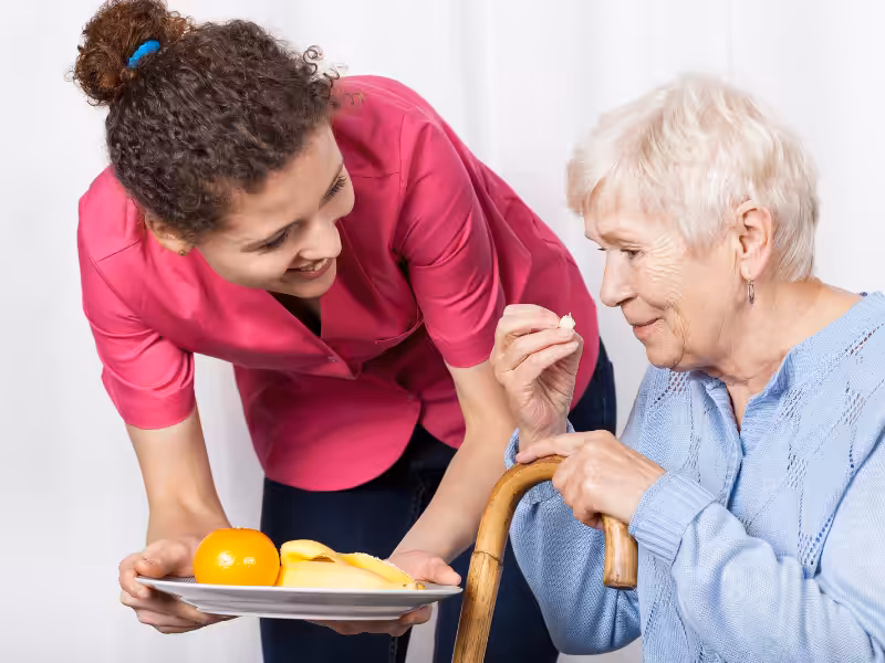 A young caregiver in a pink shirt offers a plate of fruit to an elderly woman with white hair in a blue sweater, who is holding a cane and about to take a bite of food.
