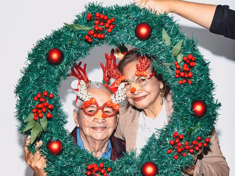 Two smiling older adults wear festive reindeer antler glasses, posing together behind a large Christmas wreath decorated with red berries and ornaments. A hand holds the wreath in front of a white background.
