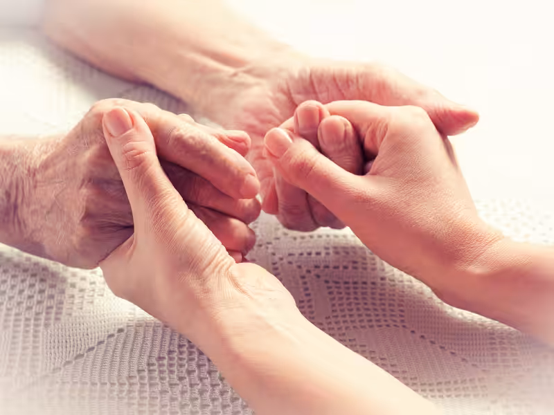 Close-up of an older person’s hands being gently held by a younger person’s hands on a white textured surface, conveying care, support, and connection.