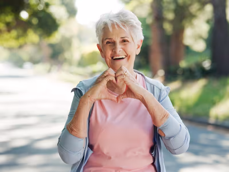 Smiling older woman with short gray hair makes a heart shape with her hands. She is wearing a light blue jacket over a pink shirt and standing outdoors on a sunny day with trees in the background.