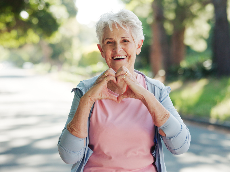 Smiling older woman with short gray hair makes a heart shape with her hands. She is wearing a light blue jacket over a pink shirt and standing outdoors on a sunny day with trees in the background.