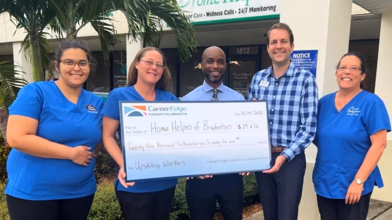 Five people stand outside holding a large check for $29,676 made out to Home Helpers of Bradenton from CareerEdge, dated 5/12/22, for “Upskilling Workers.” They are smiling and appear happy.
