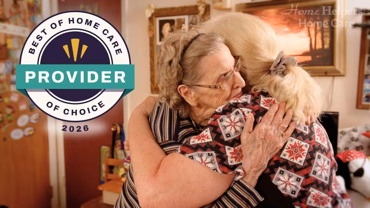 Two women warmly embrace in a cozy, decorated room. One woman is elderly with glasses, the other has blonde hair. A “Best of Home Care Provider of Choice 2026” badge is displayed on the left side of the image.