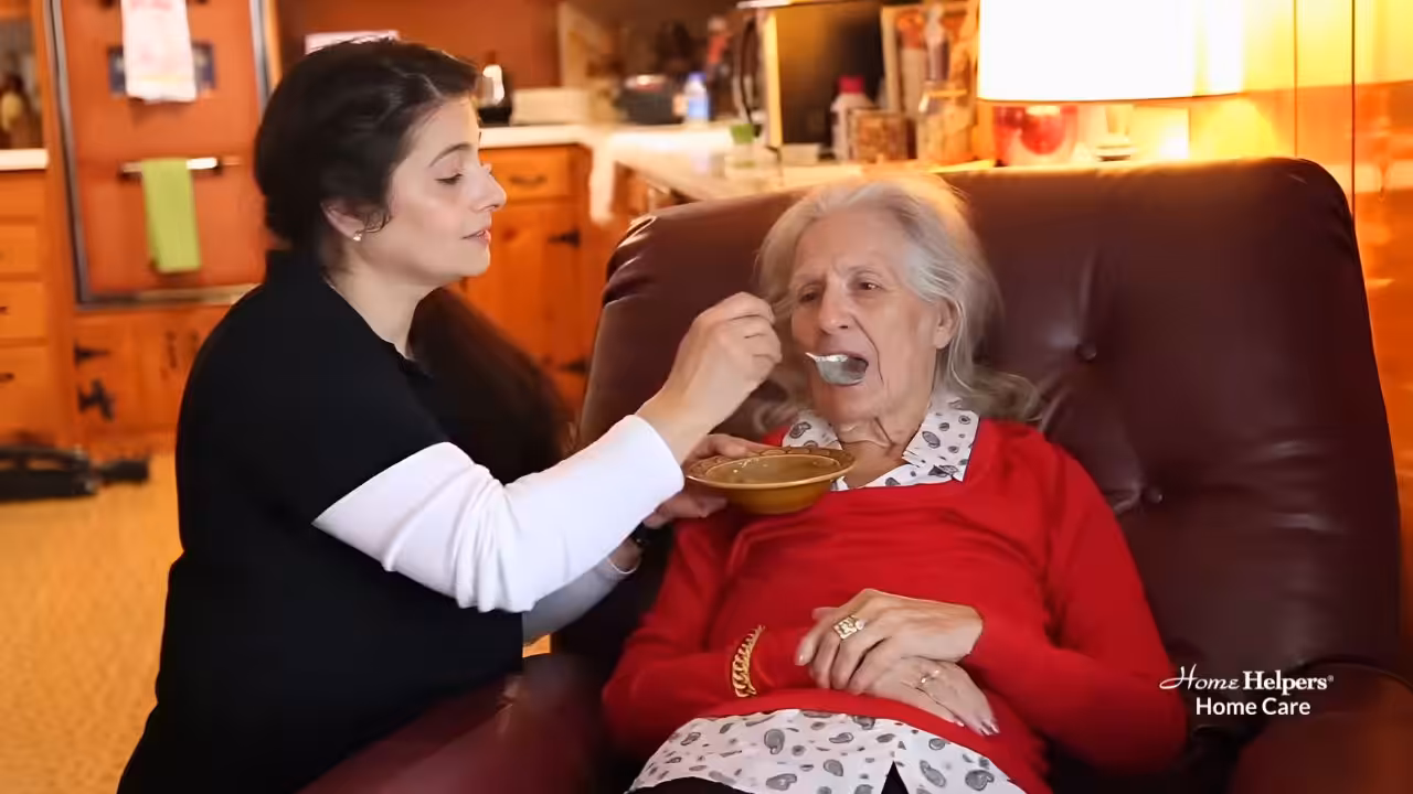 A caregiver feeds an elderly woman soup as she sits in a recliner in a cozy, wood-paneled home. The scene conveys warmth and attentive home care, with Home Helpers Home Care logo in the bottom right corner.