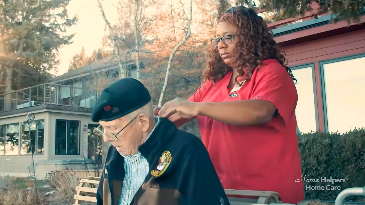 A caregiver in a red uniform helps an elderly man wearing a black beret and jacket put on a coat outdoors near a house, with trees and sunlight in the background. The words Home Helpers Home Care appear in the corner.