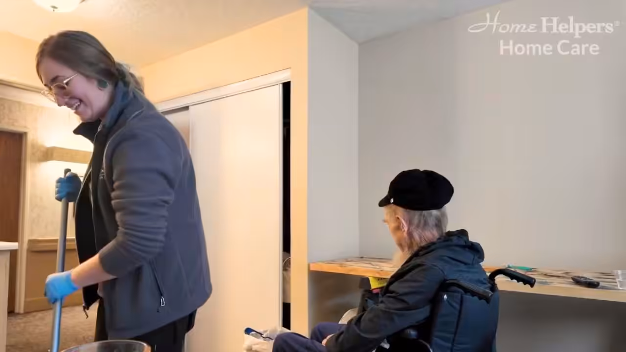 A smiling caregiver wearing gloves cleans with a mop, while an older man in a wheelchair watches. The room is bright, and Home Helpers Home Care is visible in the top right corner.