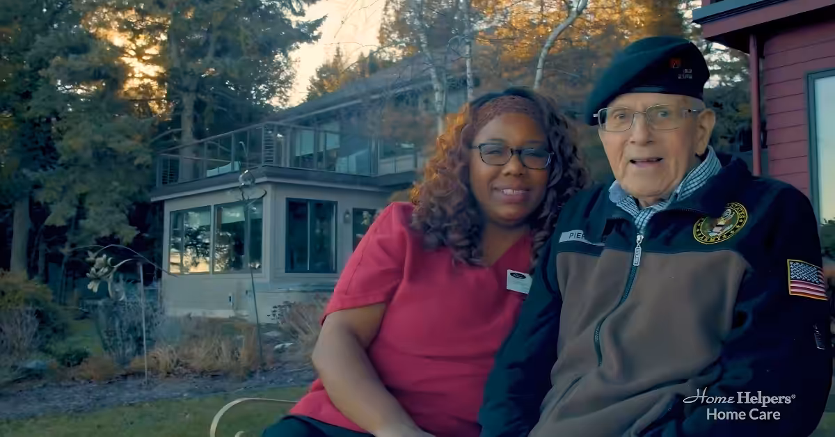 A caregiver in red scrubs sits beside an older man wearing a military-style jacket and beret. They smile outside a house with trees in the background. Home Helpers Home Care logo is visible in the corner.