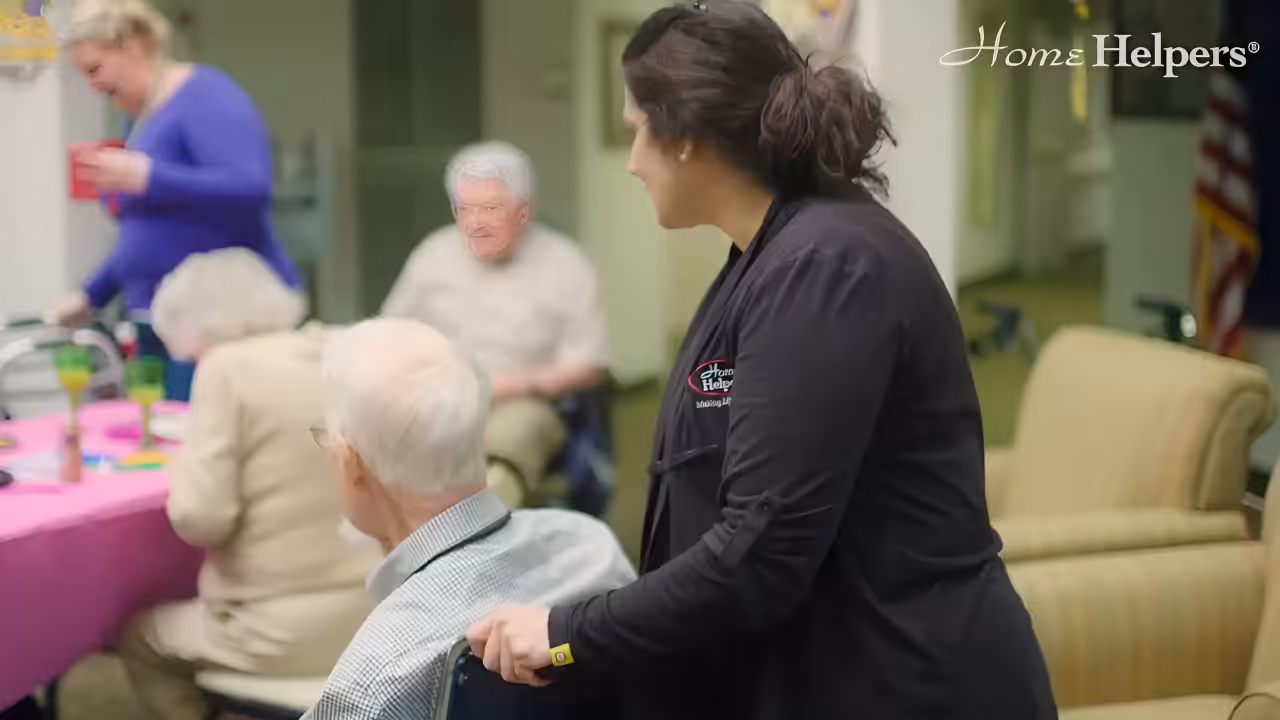 A caregiver stands beside an elderly man in a wheelchair at a community gathering. Other seniors are seated at a table with pink tablecloth in a bright, welcoming room. Home Helpers logo appears in the top right corner.
