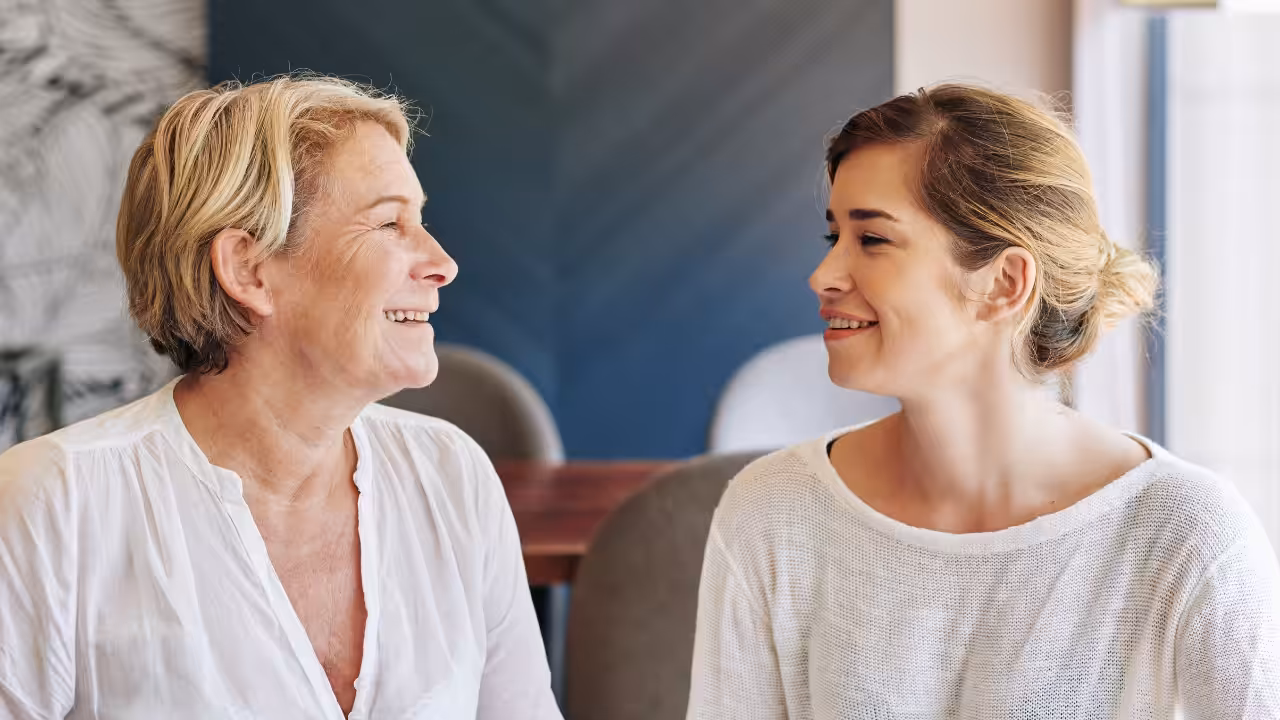 Two women, one older and one younger, sit indoors facing each other and smiling warmly. They are both wearing white tops and appear to be enjoying a pleasant conversation.
