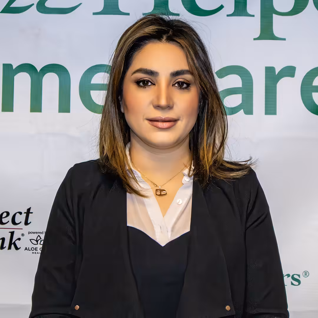 A woman with shoulder-length brown hair wearing a black blazer and white blouse stands in front of a banner with large green and black text. She is facing the camera and smiling slightly.
