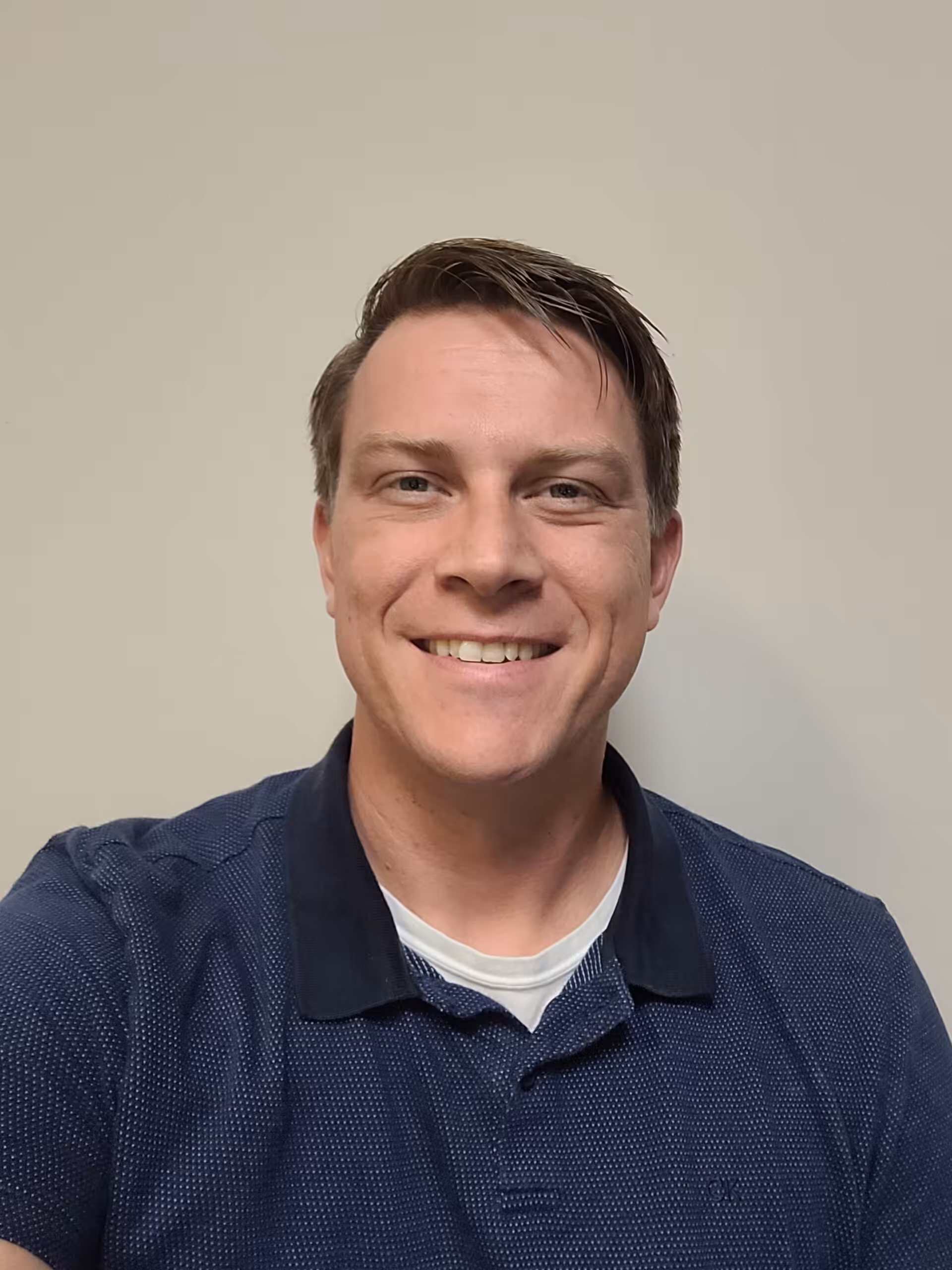 A man with short brown hair, wearing a blue collared shirt over a white undershirt, is smiling at the camera in front of a plain, light-colored wall.