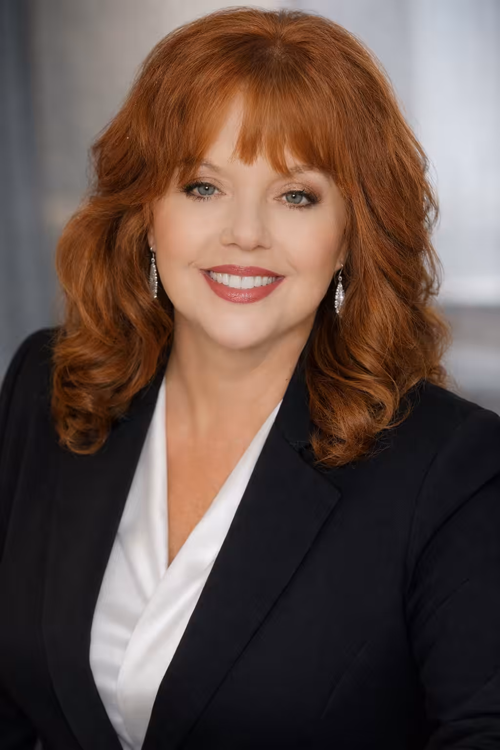 A woman with wavy red hair and light skin is smiling. She wears a black blazer, a white blouse, and dangling earrings, standing against a softly blurred background.